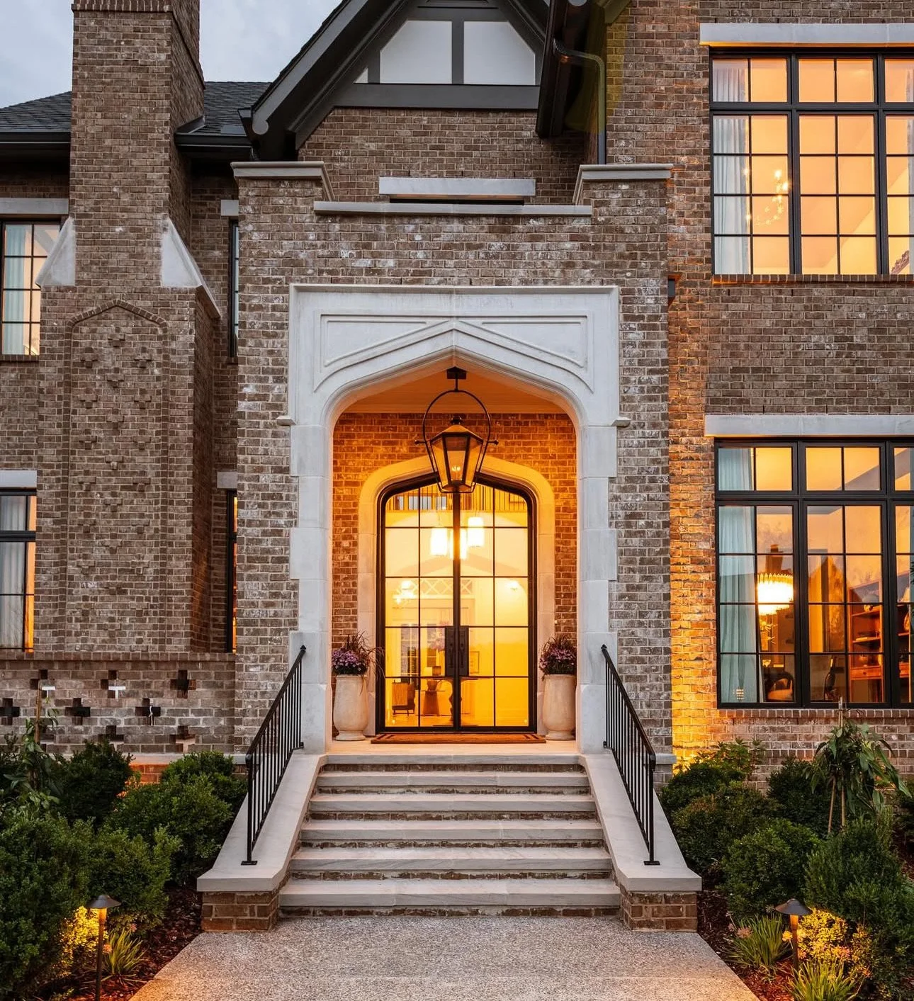 Front view of a brick house with illuminated interior, large glass front door, and steps leading up to the entrance. There are potted plants on either side of the door and landscaped bushes at the base of the stairs. The house is lit with warm lighti
