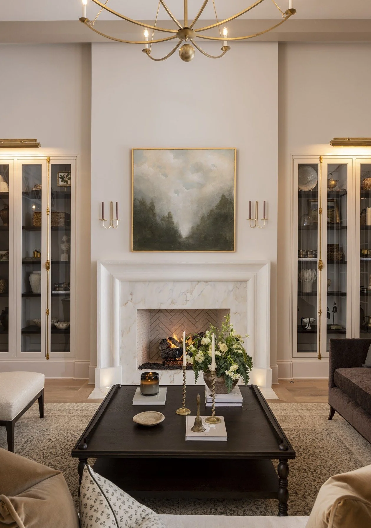 A living room with a white marble fireplace, a painting above it, and built-in glass cabinets on either side displaying decorative items. A black coffee table with candles, books, and plants is in the center, with sofas and an area rug around it. A g
