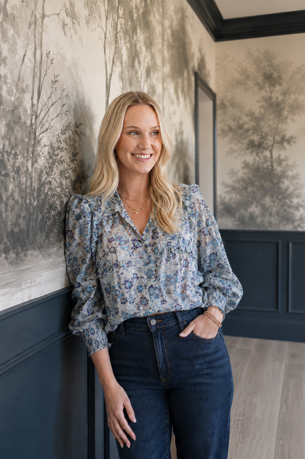A woman with blonde hair, wearing a patterned blouse and dark jeans, leaning against a wall with a scenic forest mural, smiling and looking to the side.
