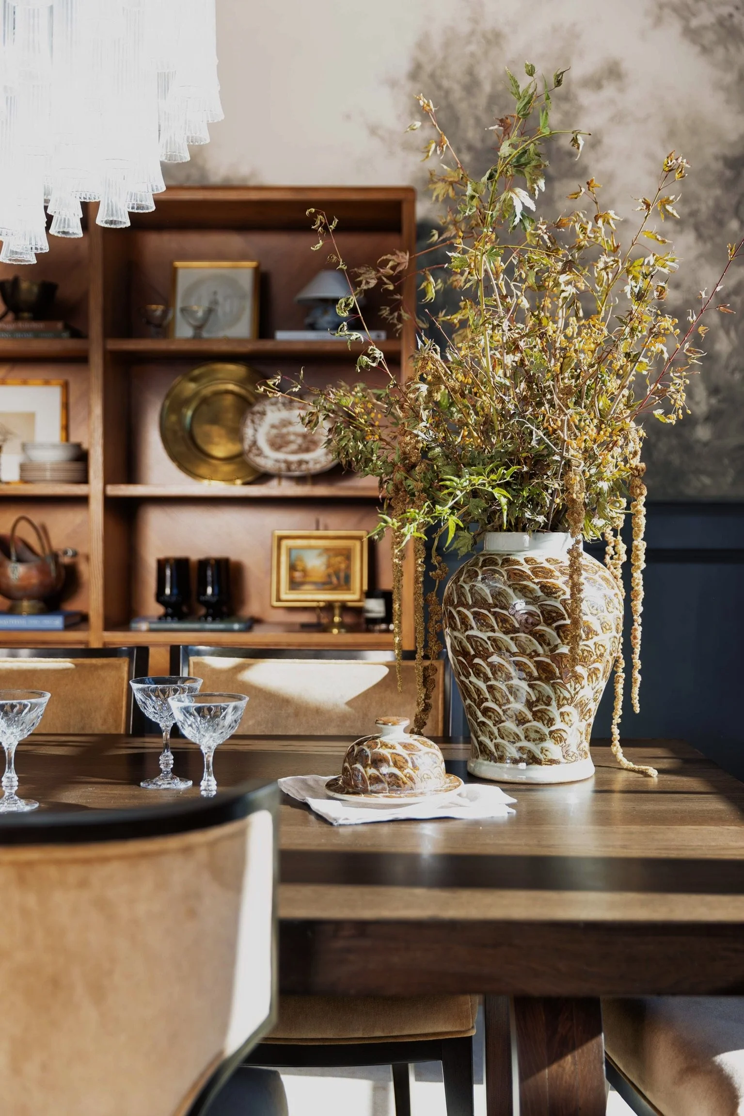 A decorated dining table with a large ornate vase filled with dried and withered flowers, crystal glasses, and a small serving dish with a lid. In the background, a wooden bookshelf displays framed art, gold and ceramic plates, bowls, and decorative items, with a textured wall behind.