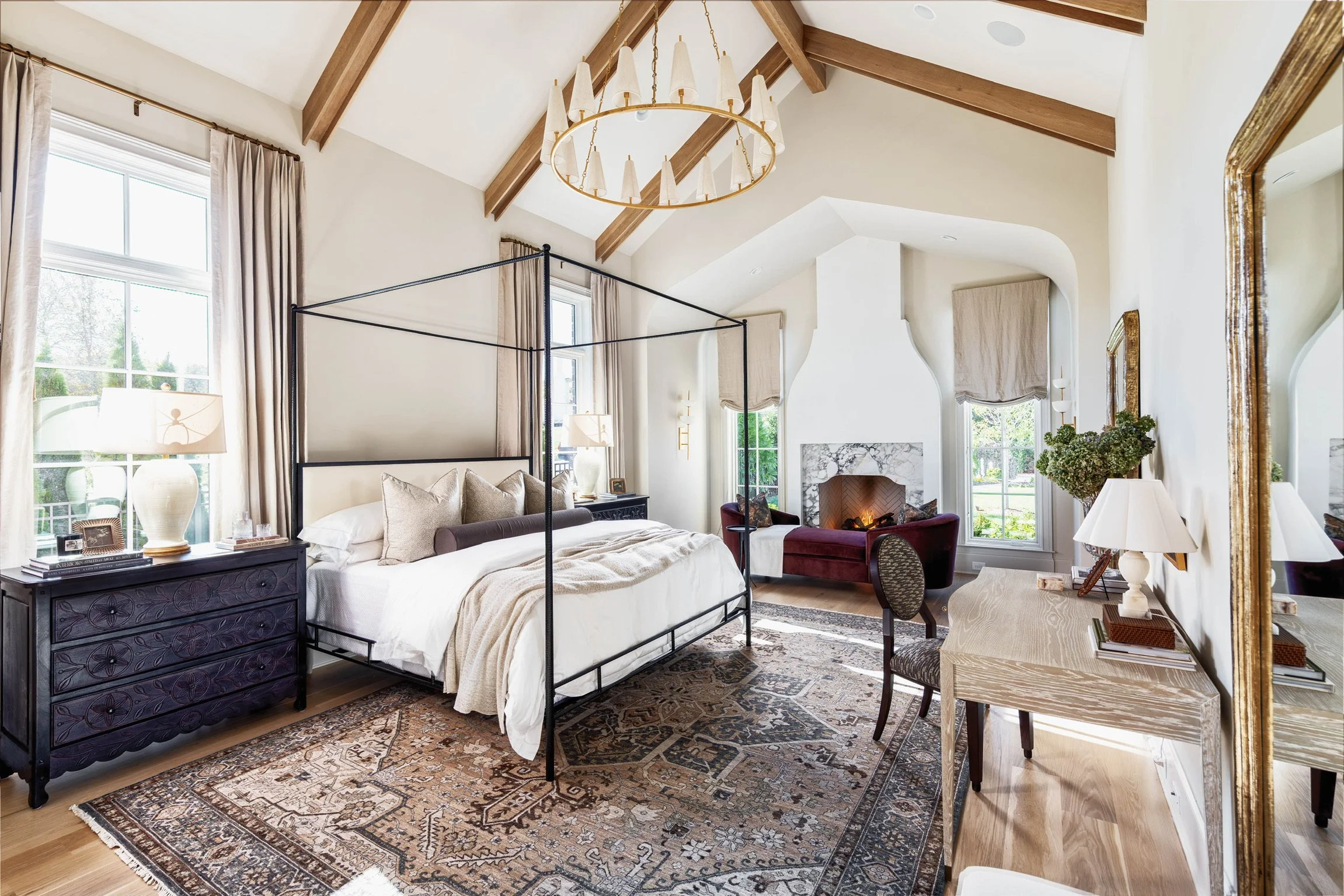 A spacious bedroom with high vaulted ceiling featuring wooden beams and a modern chandelier. The room has large windows with beige curtains, a black metal canopy bed with white bedding, a dark wooden dresser, a fireplace with a marble surround, two a