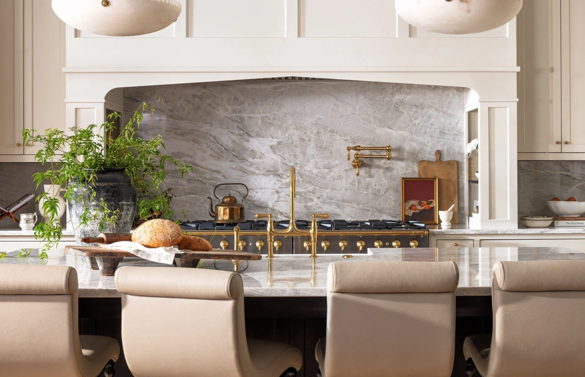 A kitchen countertop with a green potted plant, bread, and a black tray. Behind it is a stovetop with brass fixtures, a granite backsplash, and white cabinetry.