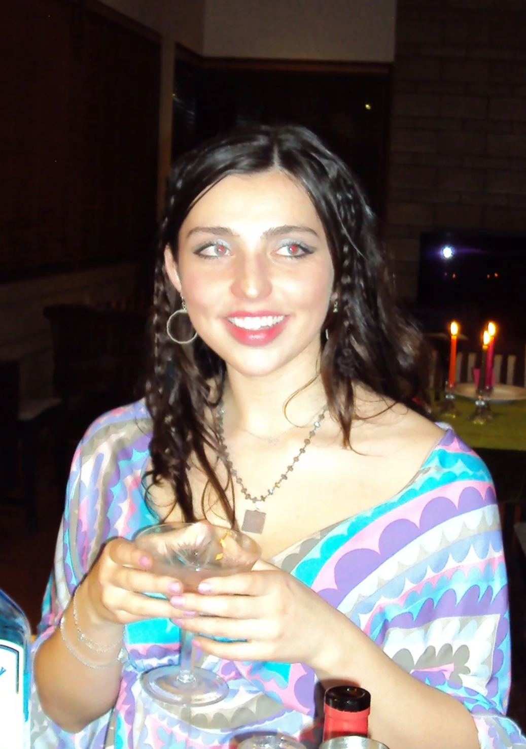 A woman with wavy dark hair, wearing hoop earrings, a necklace, and a colorful patterned dress, smiling and holding a glass of wine spritzer (Maison Maxine) at a party or gathering in a dimly lit room.