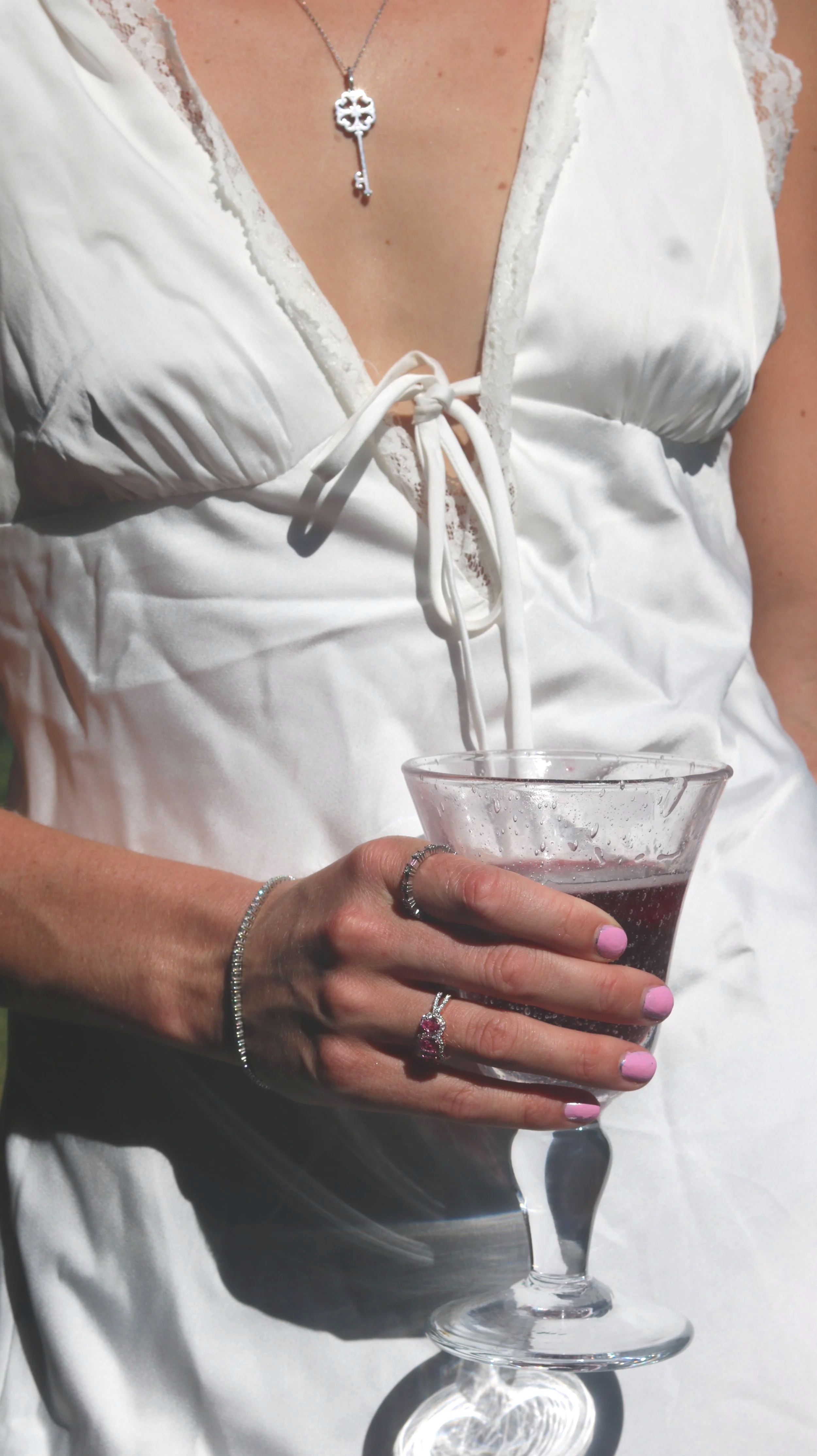 A woman in a white dress holds a martini glass with a pink beverage. She is wearing jewelry, including rings, a bracelet, and a necklace with a key pendant.