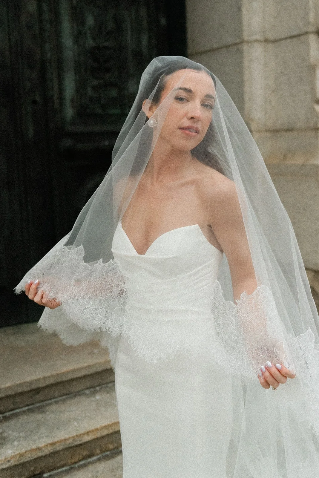 A woman in a white wedding dress with lace accents at the sleeves and a veil over her face, standing outside on the steps of a building.