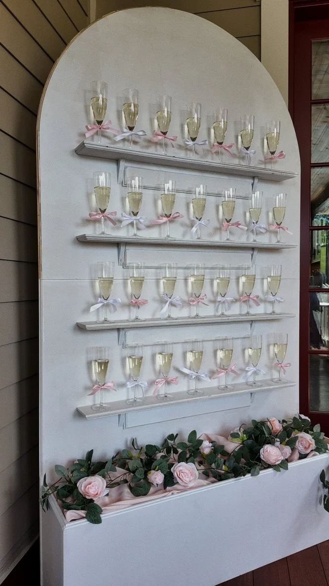 A stunning champagne glass display dressed in pink and white bows, nestled atop a rose-adorned tiered board, the kind of showstopping detail Palm Events & Rentals creates to wow guests at any wedding or event.