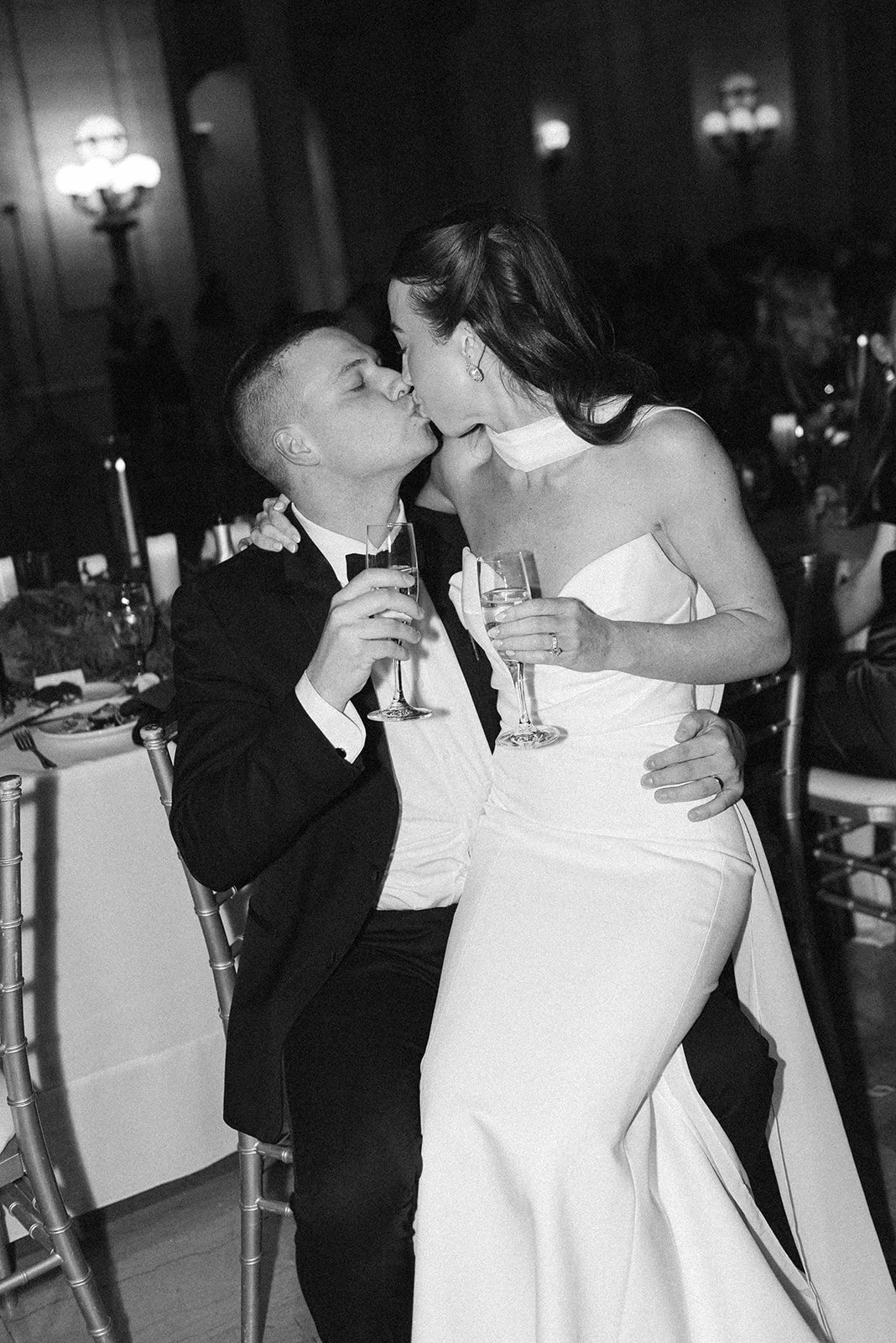 A couple dressed in wedding attire sharing a kiss at their wedding reception, holding glasses of champagne.