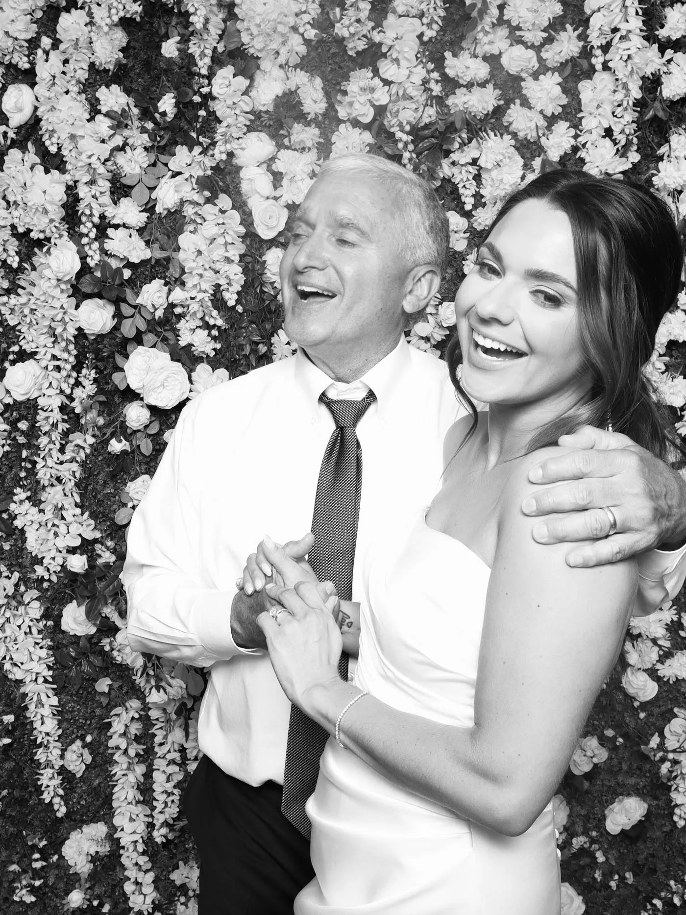 A father and daughter pose for the camera, while dancing and smiling in front of a flower wall.