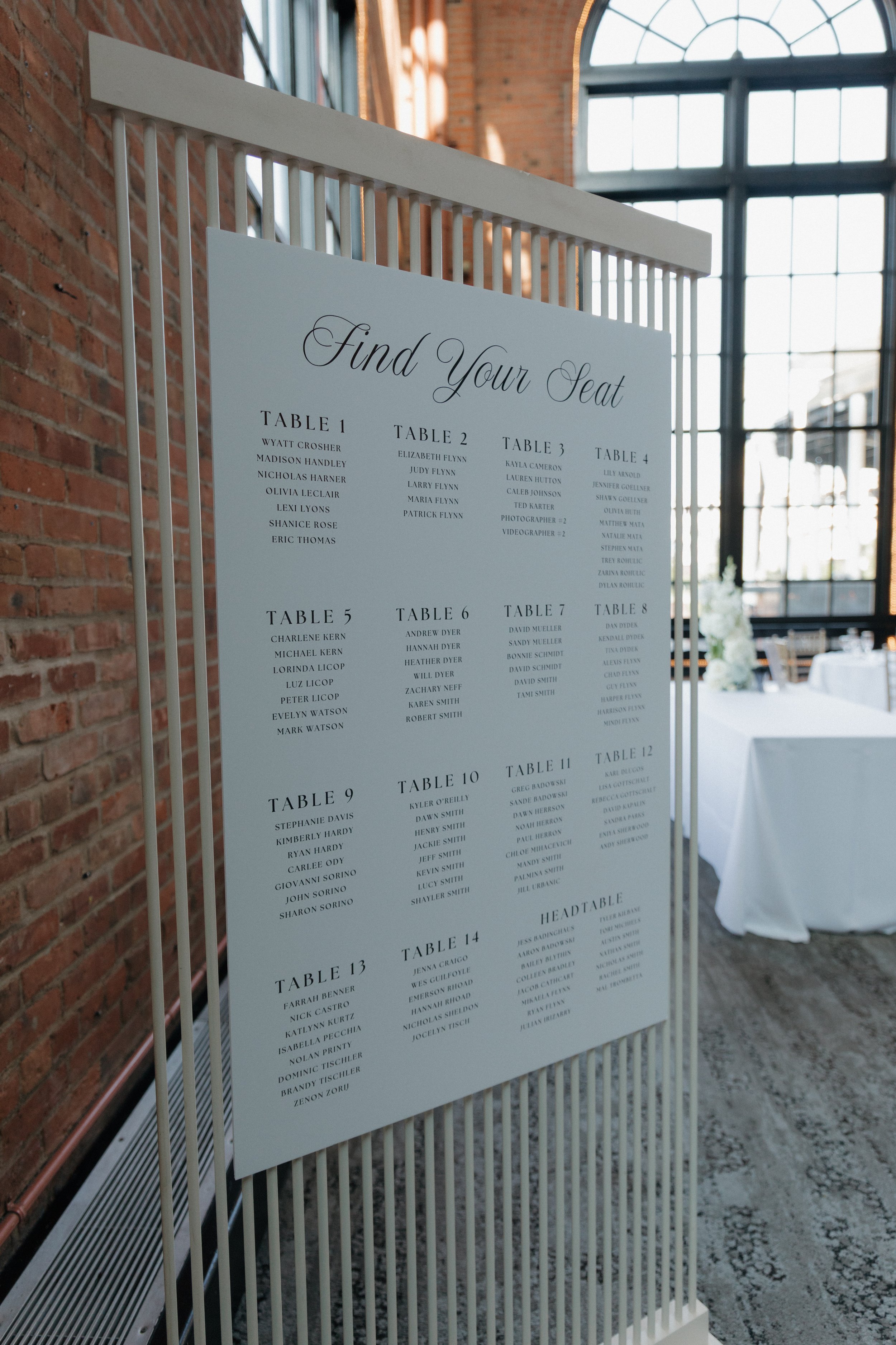 Seating chart for an event with tables numbered 1 to 14, listing attendee names under each table, titled 'Find Your Seat', displayed on a white standing frame inside a venue with exposed brick walls and large arched windows.