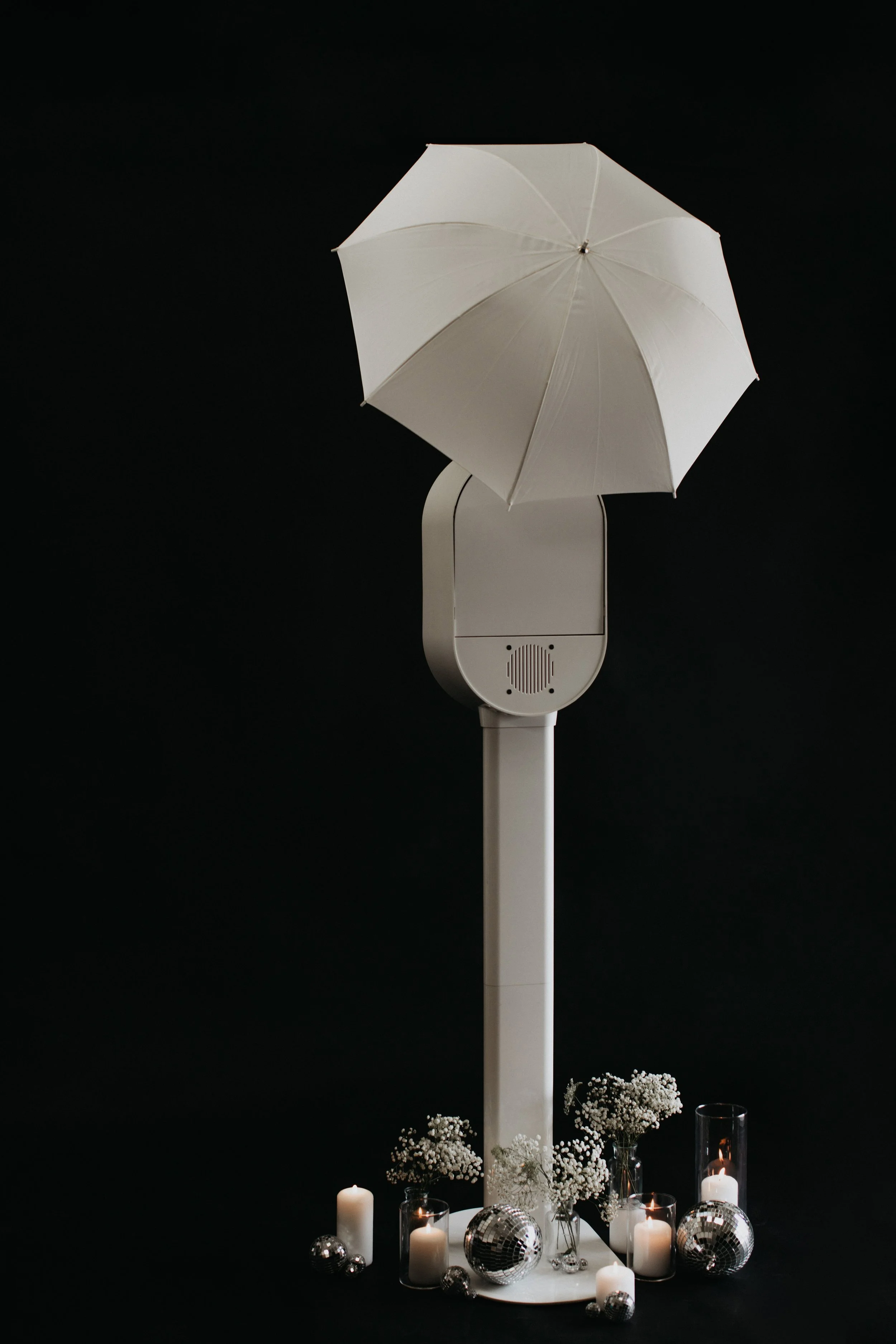 A white light modifier propped open above a modern photo booth, with candles, disco balls, and flowers arranged at the base against a dark background.