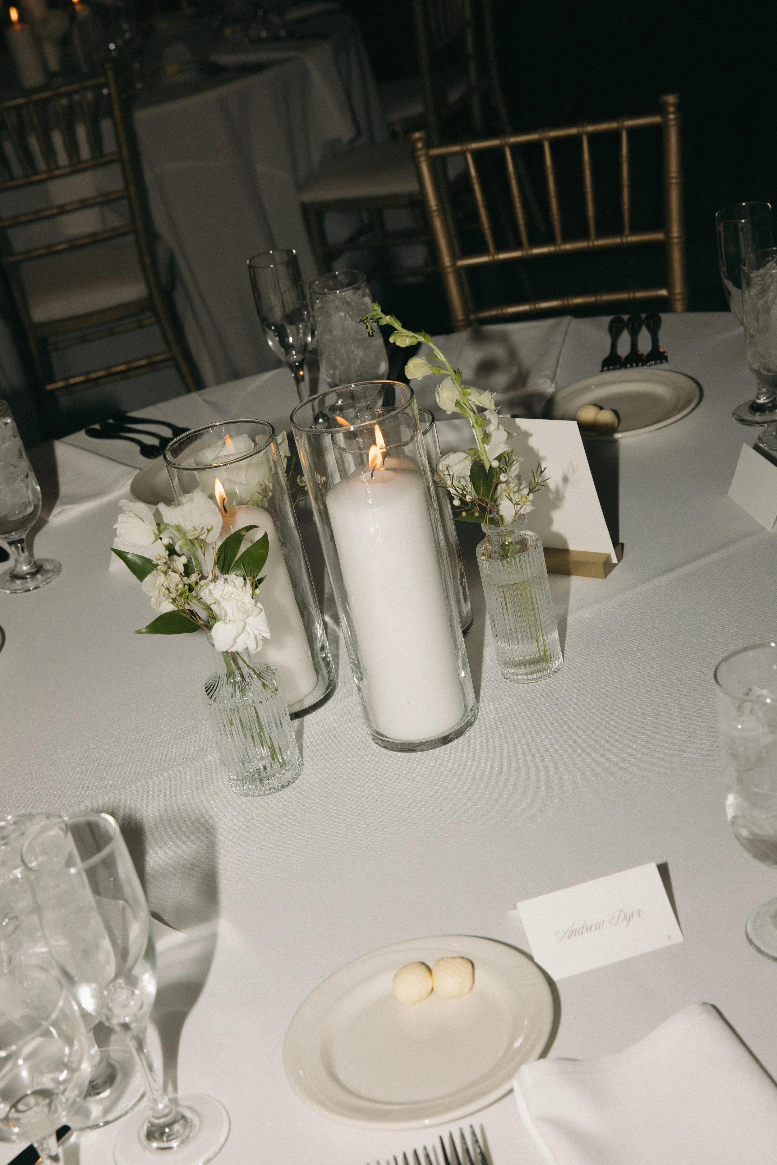 A softly styled wedding banquet table with tall glass cylinder candles, delicate white florals, and gold chairs, exactly the kind of refined design detail Palm Events & Rentals brings to every event.