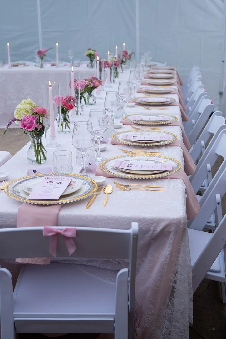 Elegant wedding or formal dinner table setup with pink and white floral centerpieces, lit candles, and pink napkins, set with gold-accented plates, gold utensils, and wine glasses.