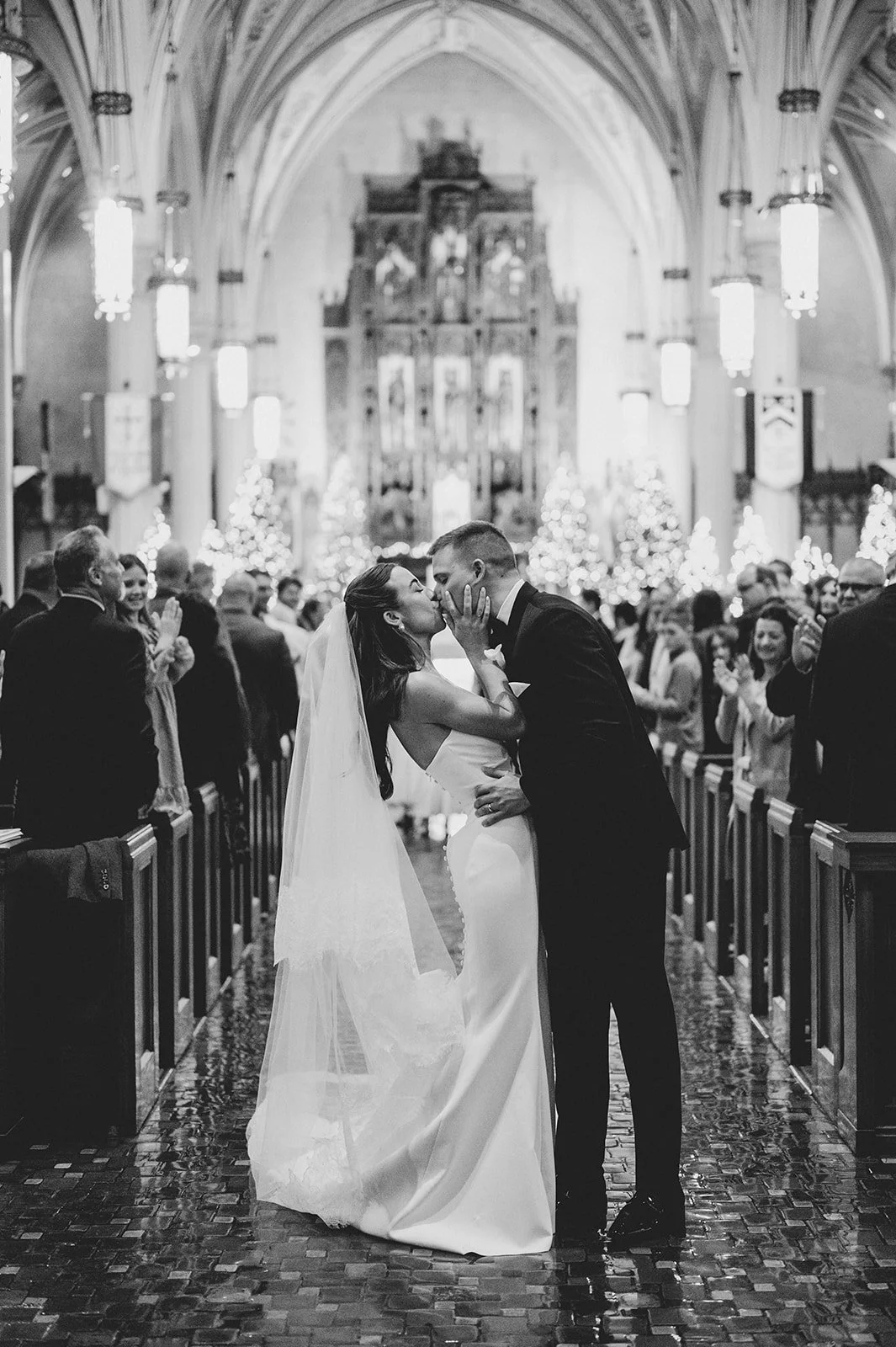 A newlywed couple sharing their first kiss at the altar of a grand church, guests lining the aisle around them, captured in moody black and white, the kind of unforgettable ceremony moment Palm Events & Rentals helps bring together.
