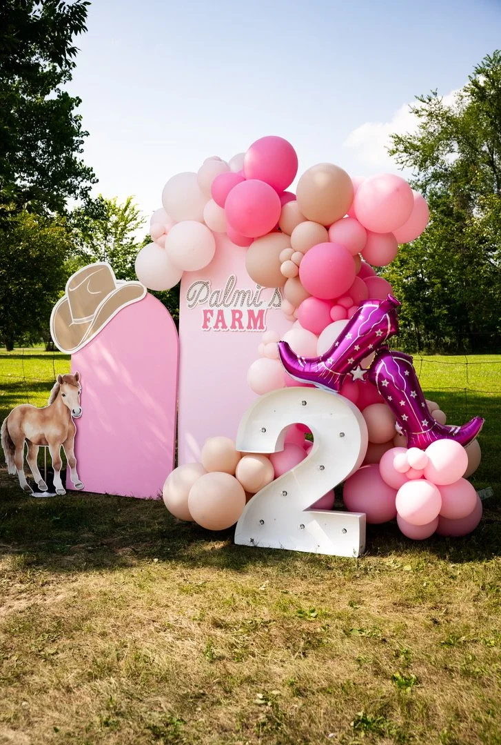 A whimsical farm-themed birthday setup with a balloon arch in pink, beige, and white, a giant number 2, and adorable cowgirl boot props, because Palm Events & Rentals brings the same magic to kids' celebrations as they do to every event.