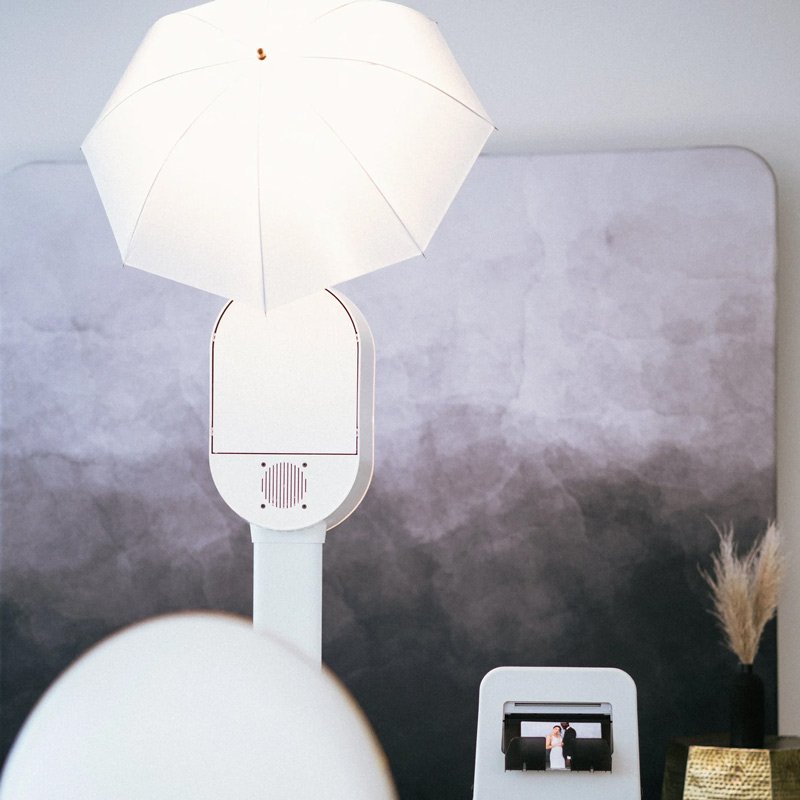 A modern photo booth with a white umbrella light modifier and dedicated printing station, styled and ready to go, the kind of polished setup Palm Events & Rentals brings to events so guests leave with beautiful keepsakes in hand.