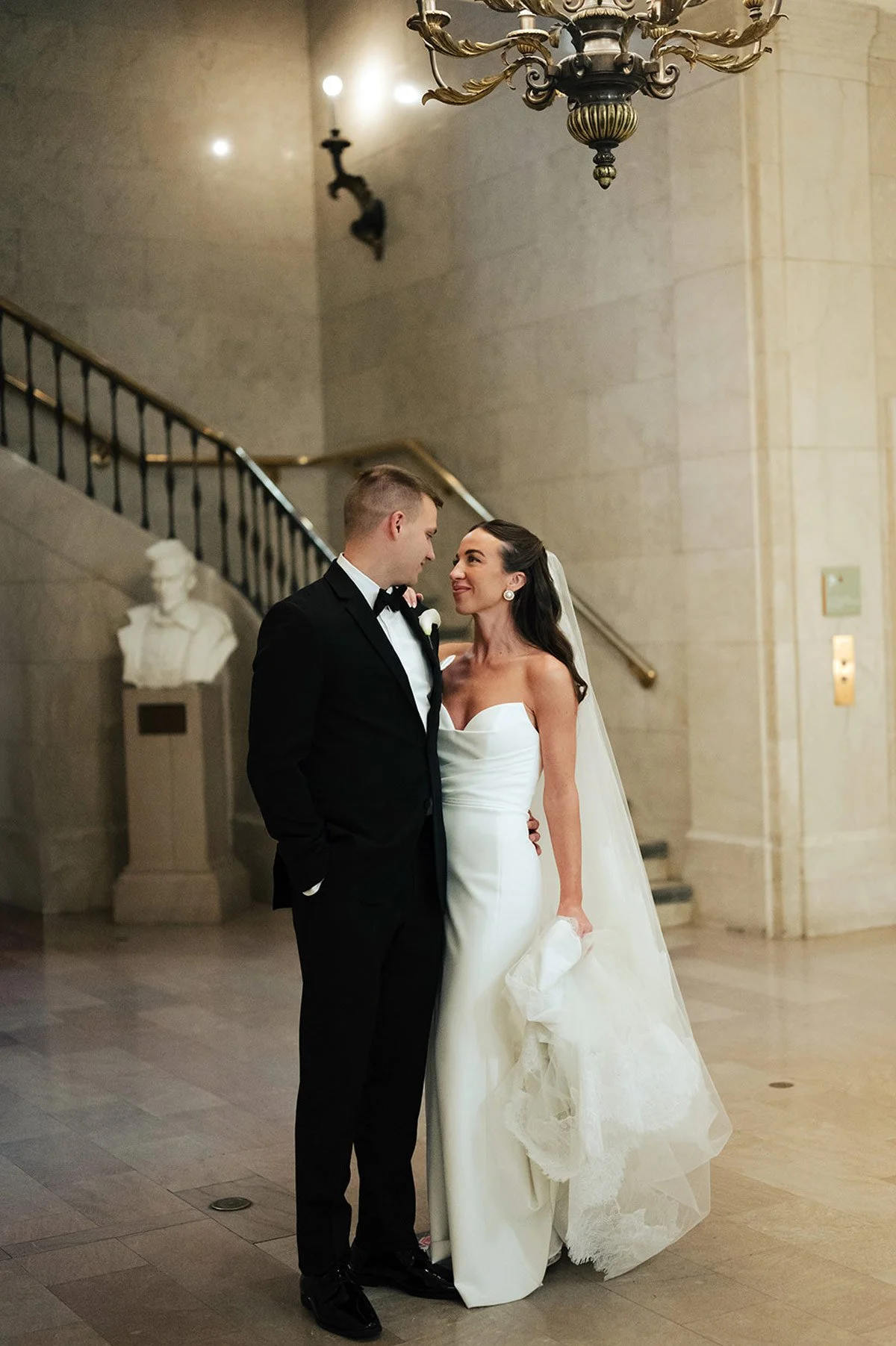A bride and groom sharing a quiet moment in a grand hall, chandelier glowing above and a sweeping staircase behind them, exactly the kind of magical wedding memory Palm Events & Rentals helps bring to life.