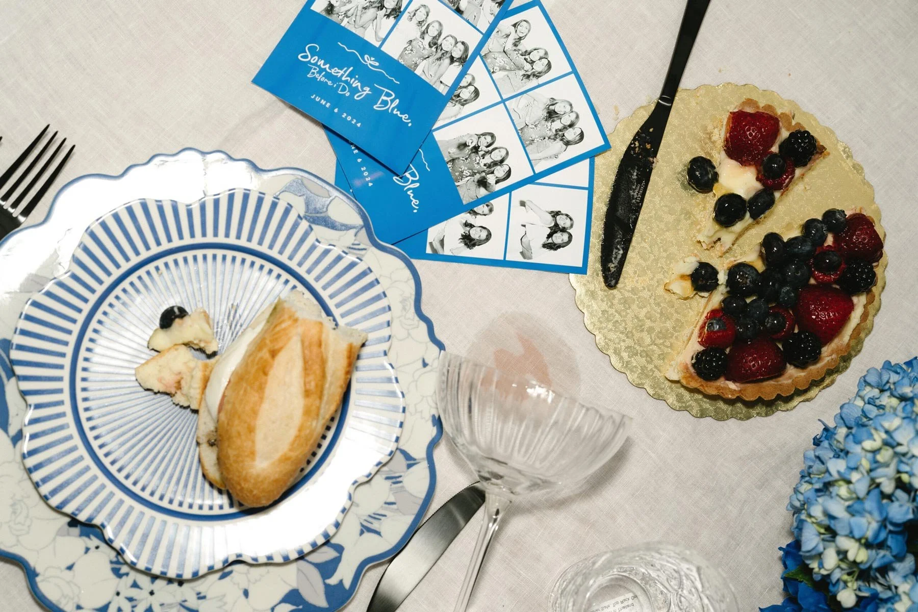 A dreamy afternoon tea flat-lay featuring a blue-striped china plate with a buttery croissant, a berry-topped tart on a gold platter, crystal glassware, and scattered Hunting Day cards on white linen.
