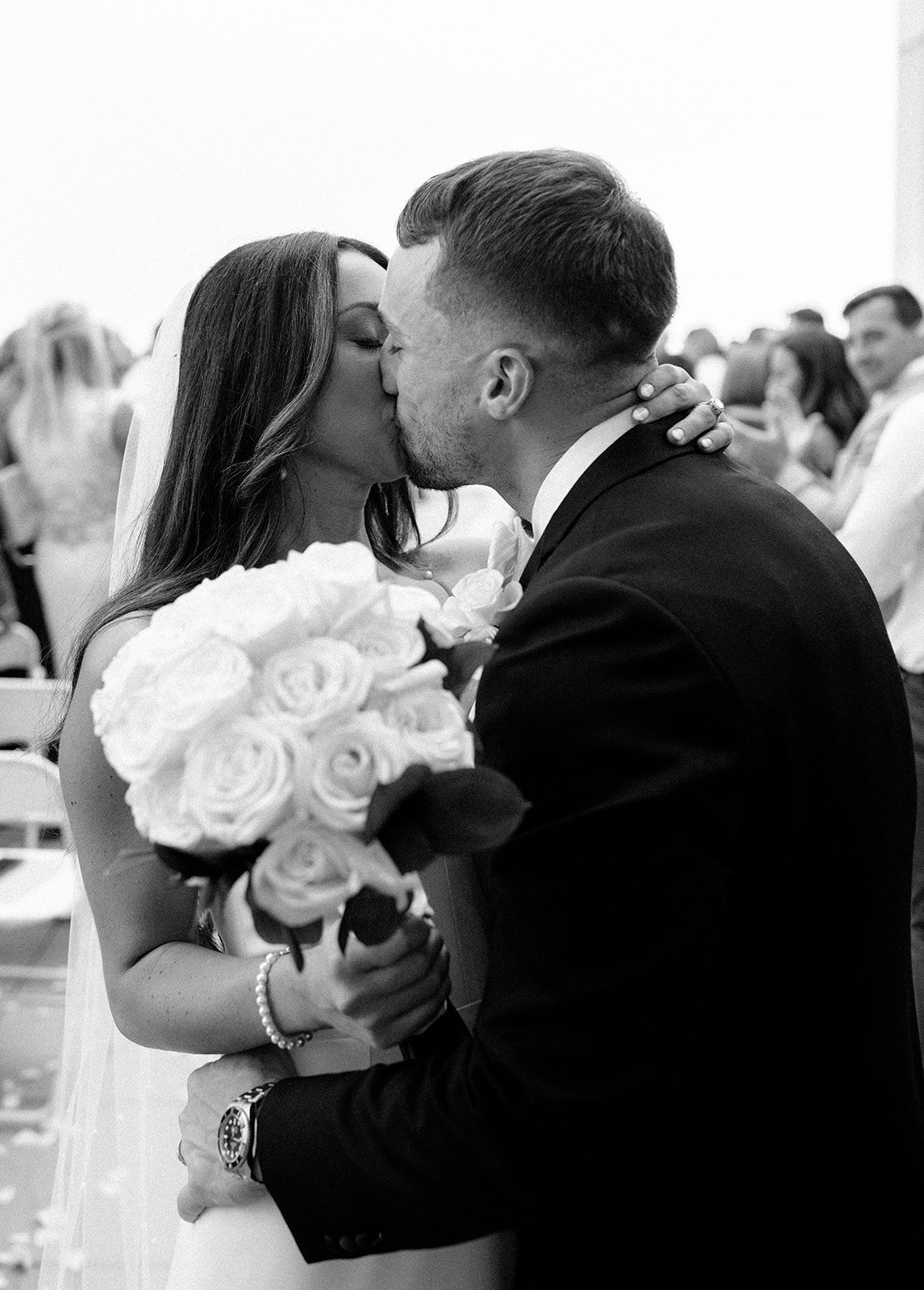 A black and white photo of a bride and groom kissing during their wedding ceremony. The bride holds a bouquet of roses, and they are surrounded by attendees.