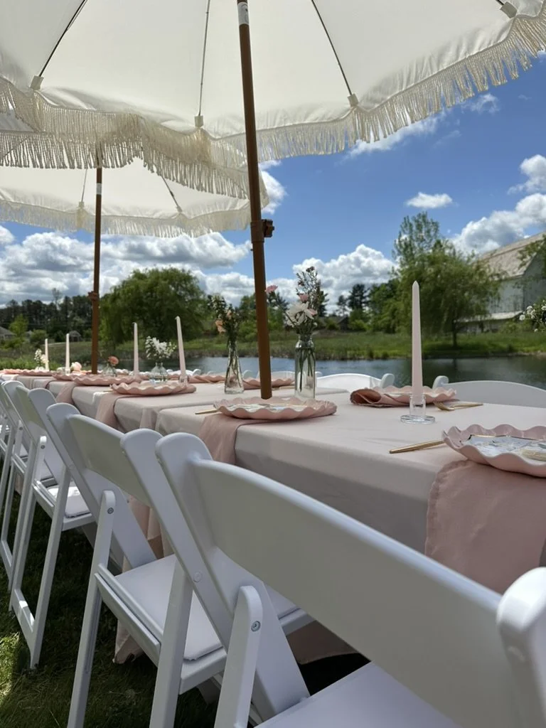 A dreamy outdoor tablescape with pink scalloped plates, white candles, and fresh florals shaded by oversized white umbrellas, with a river and blue sky as the backdrop, proof that Palm Events & Rentals creates stunning setups for indoor and outdoor e