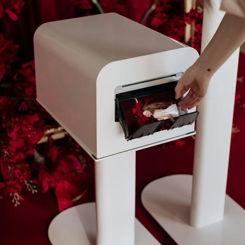 A guest pulling a fresh printed photo from a sleek white printing station, red florals glowing in the background, the kind of tangible, take-home memory Palm Events & Rentals photo booth setups deliver at every wedding and event.
