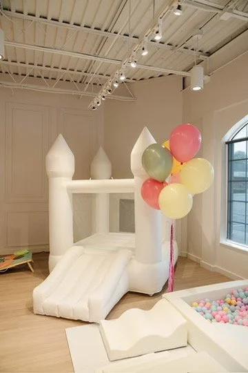 A dreamy white inflatable castle slide, pastel balloon column, and colorful ball pit set up in a bright airy space, exactly the kind of kid-approved entertainment Palm Events & Rentals provides for children's parties and family events.