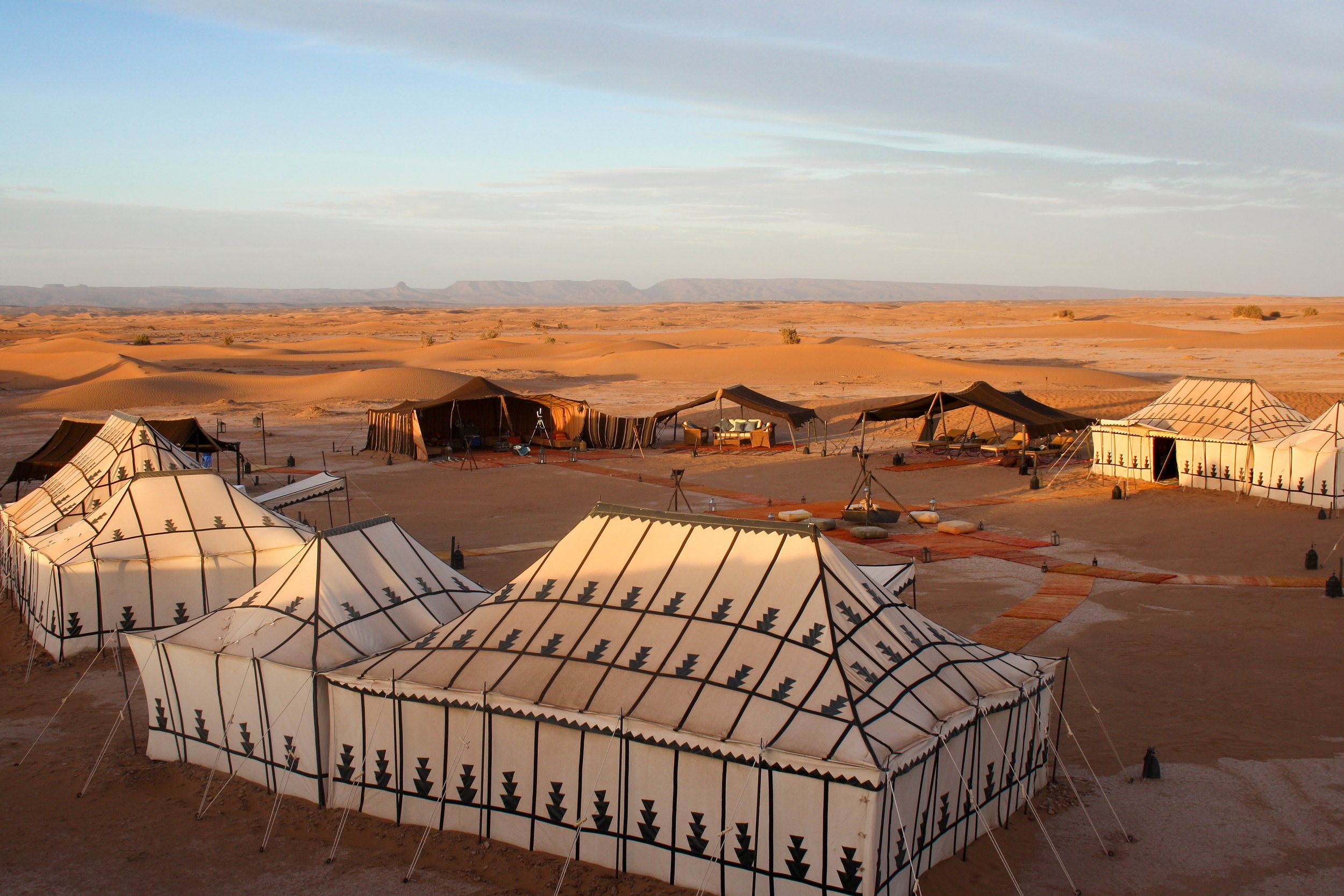 A desert landscape with sand dunes and a bivouac camp featuring tents and shade structures, under a partly cloudy sky.