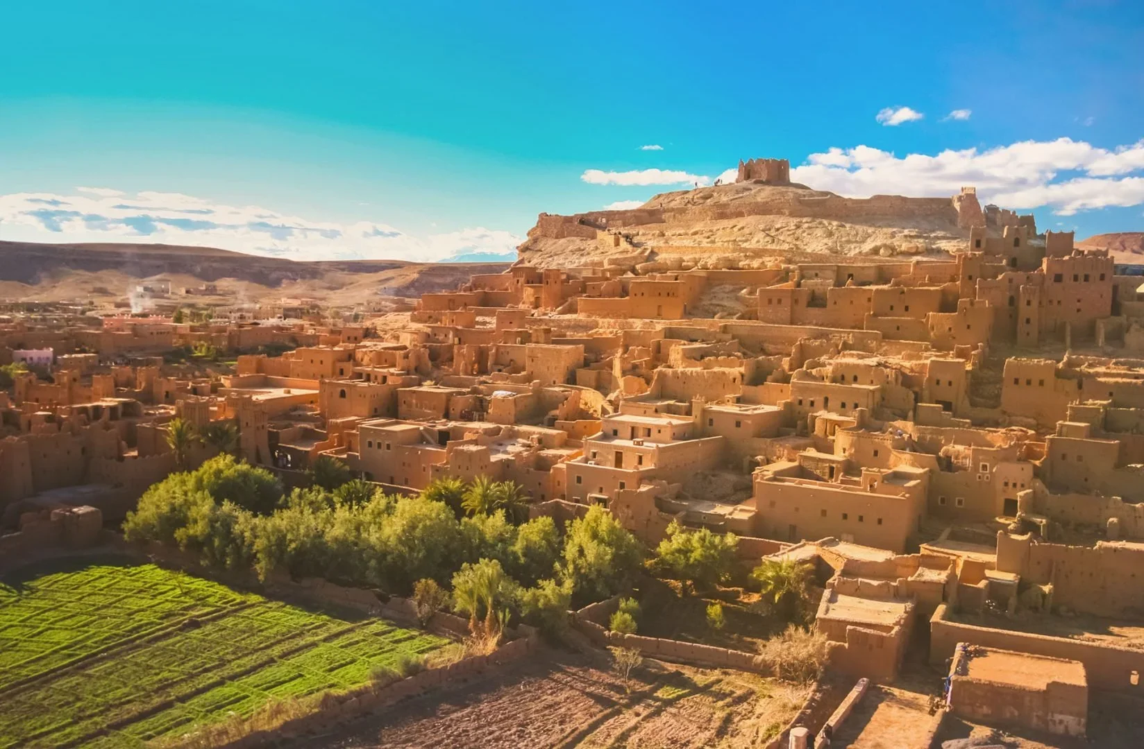 A desert town with numerous adobe-style buildings on a hillside, overlooking lush green fields with trees and mountains in the background under a blue sky with scattered clouds.