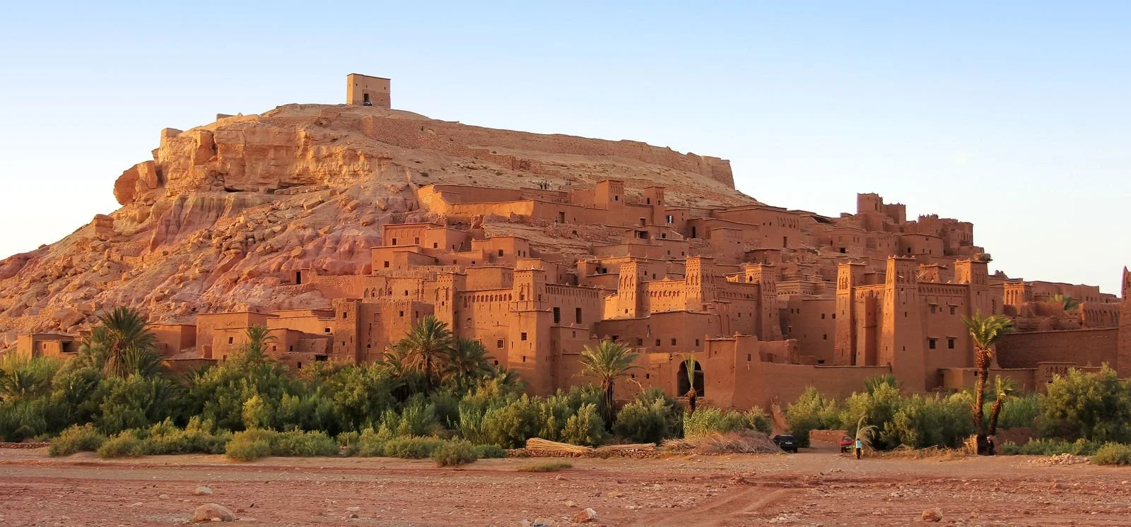 Ancient settlement built into a desert hillside with adobe-style buildings and palm trees in the foreground.