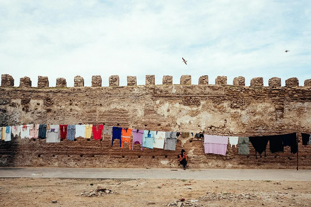 Laundry hanging on a line below a stone wall, with birds flying above and a person sitting on the ground nearby.