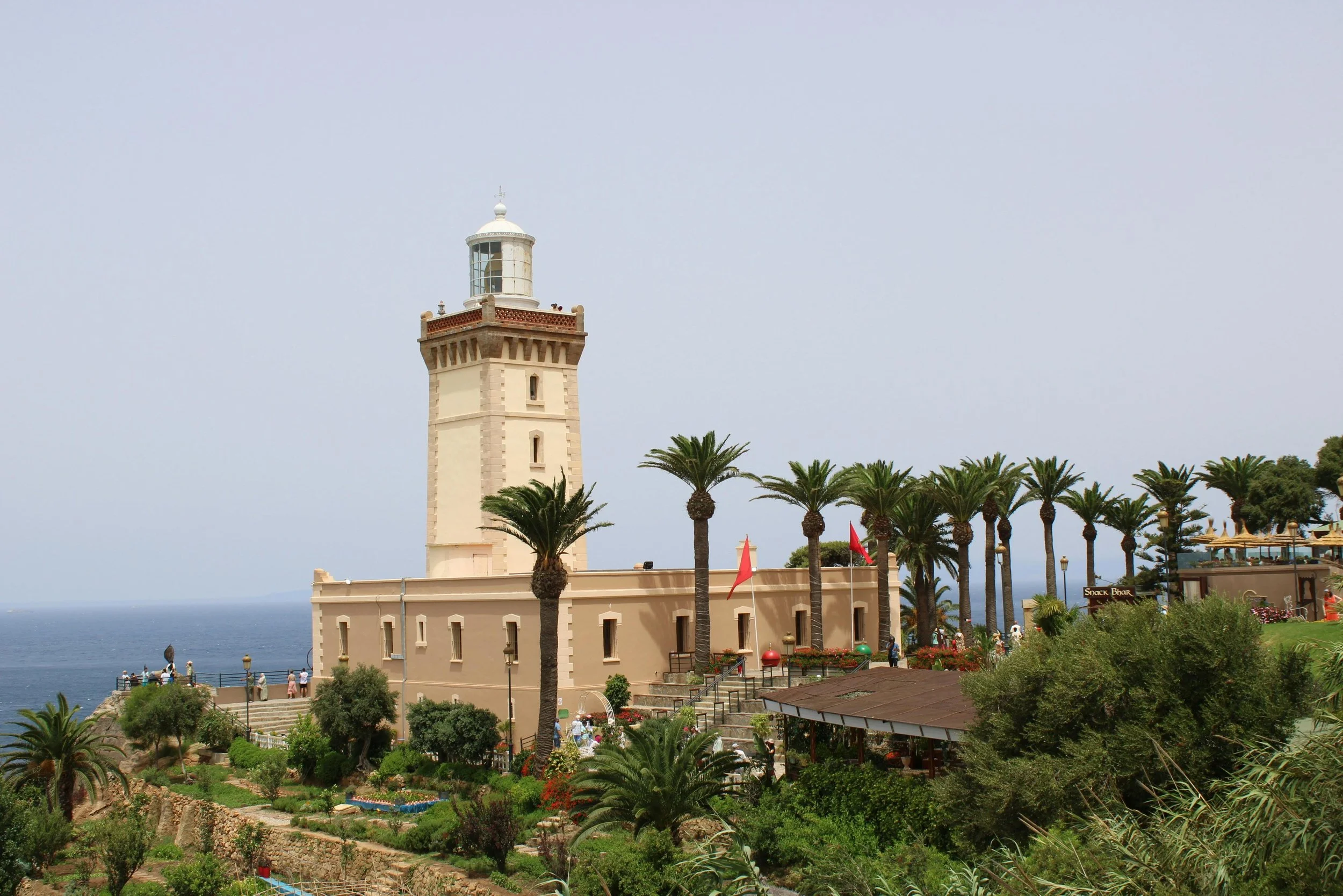 A scenic view of a lighthouse with a white tower and lantern room, surrounded by palm trees, with the ocean in the background and a partly cloudy sky.