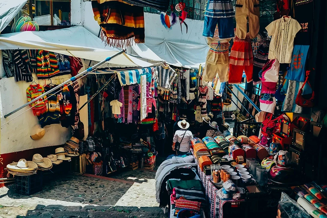An outdoor market stall displaying colorful traditional clothing, hats, woven bags, and accessories under a white canopy. People are browsing the items.
