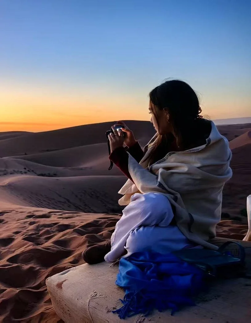 Woman sitting on a sand dune in a desert at sunset, taking a photo with a camera that she is holding in both hands