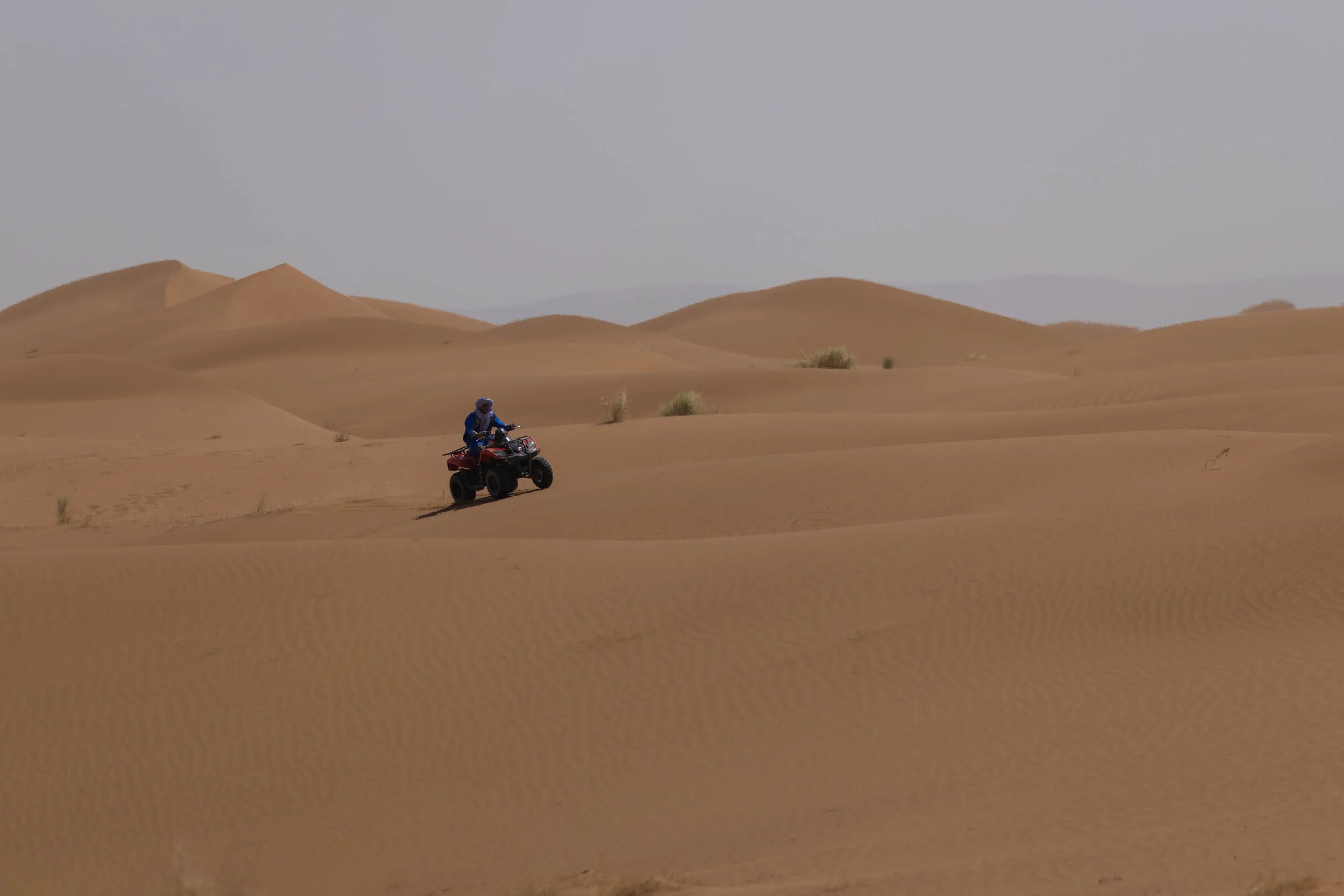 A person riding a four-wheeler vehicle across sandy dunes in a desert landscape.