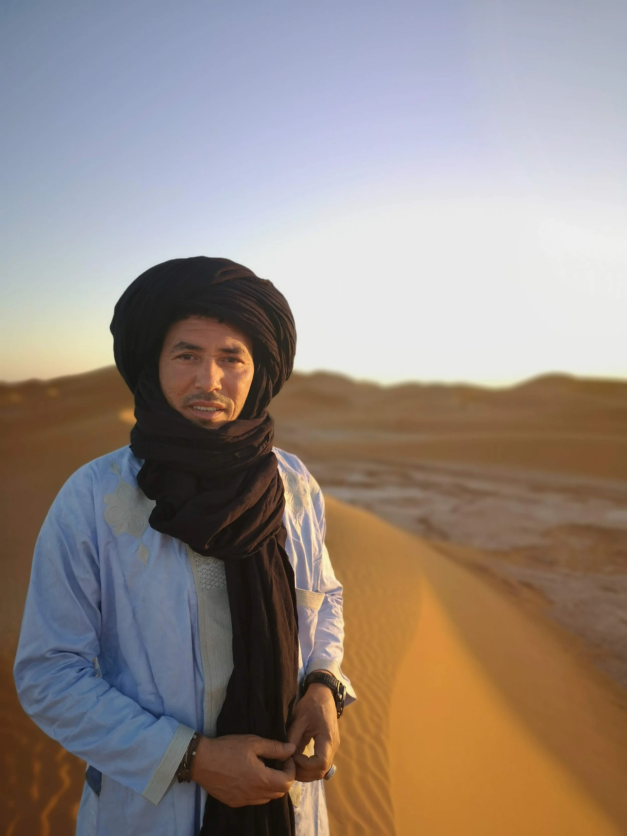 A man wearing traditional desert attire, including a large black turban and light-colored shirt, standing in a sandy desert landscape during sunset.