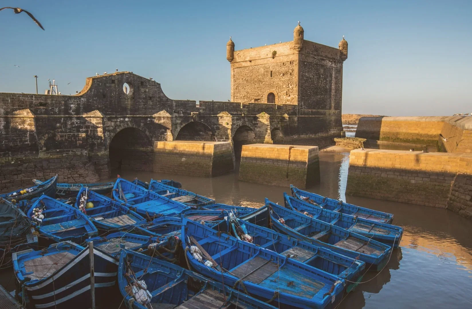Historic stone fort with rounded towers and archways by the water, with several blue fishing boats docked in the foreground during the daytime.