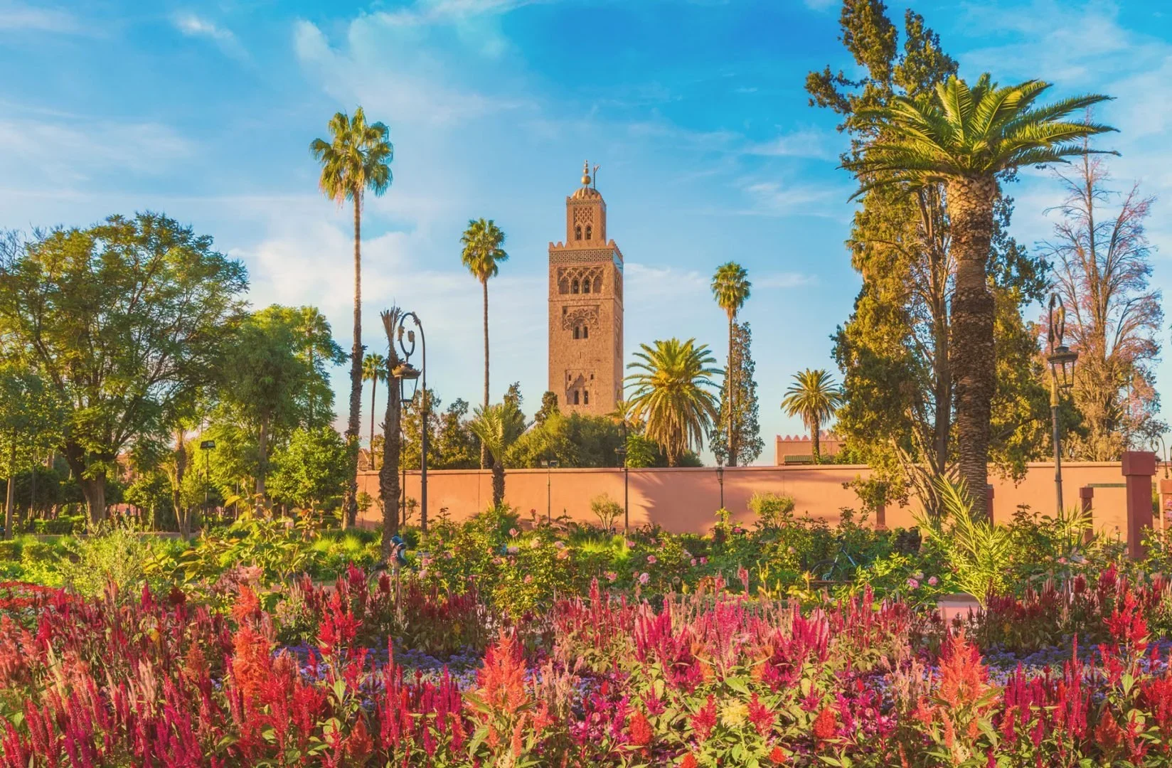 A tall clock tower surrounded by palm trees and lush greenery in a garden with vibrant pink, purple, and red flowers under a blue sky with scattered clouds.