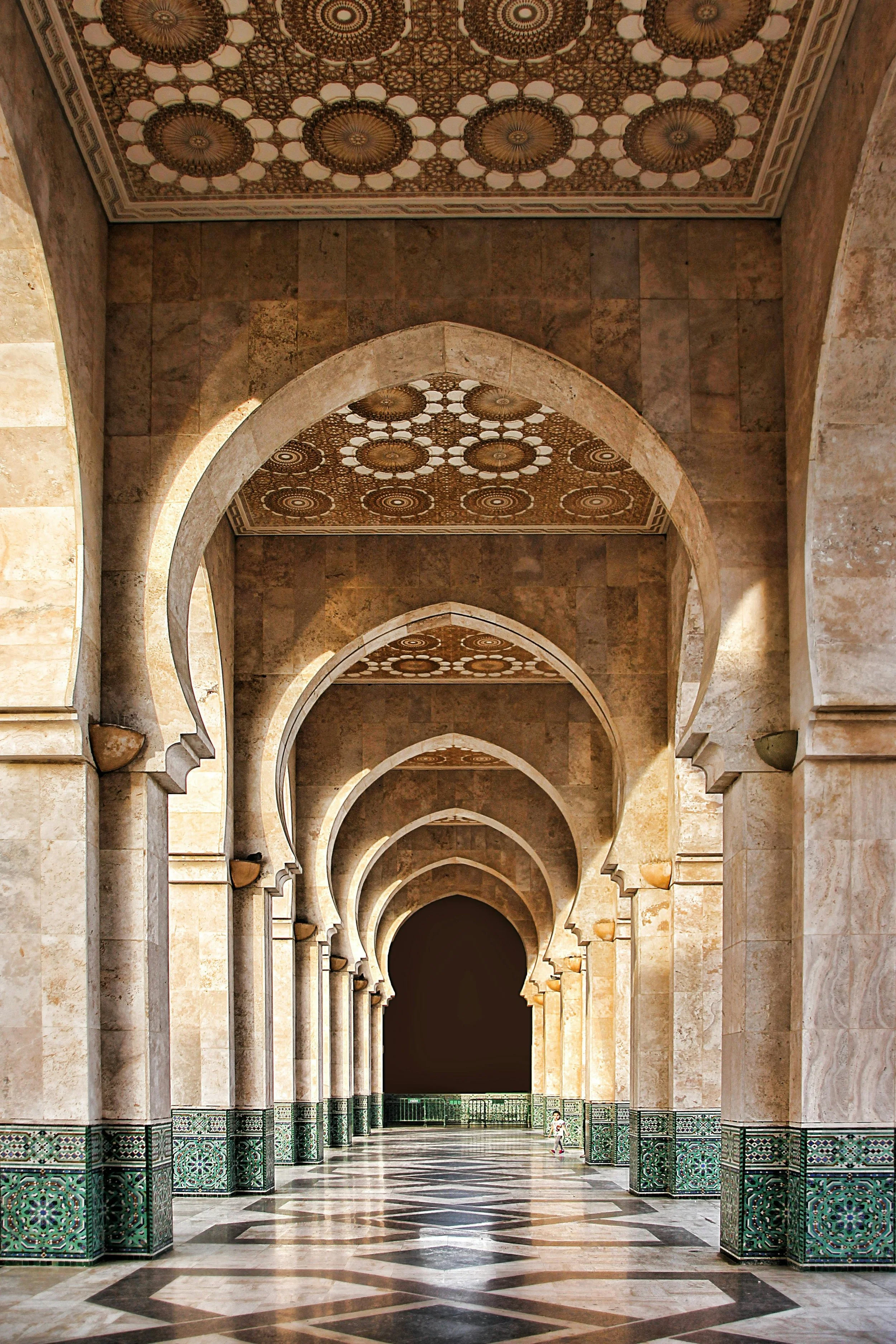 Grand corridor with arches, intricate ceiling designs, stone pillars, and tiled floor, featuring a small child walking in the distance.