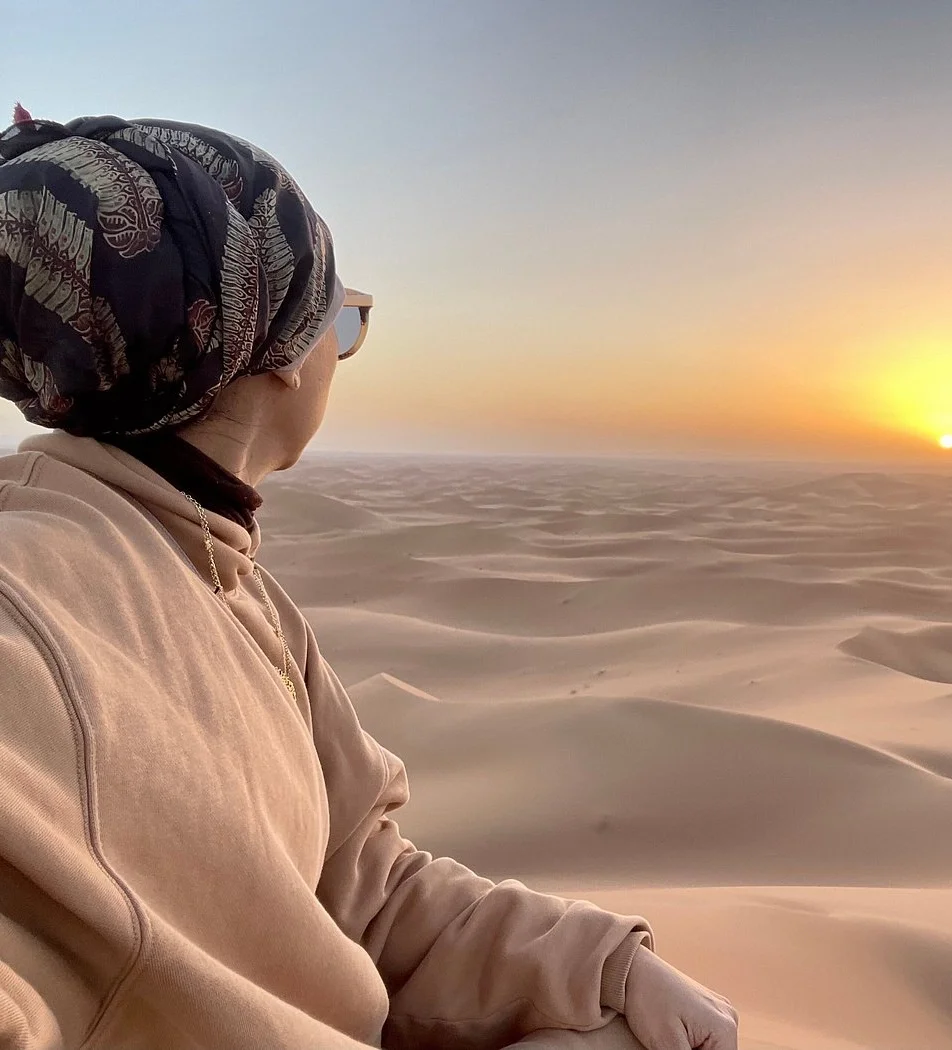 A person sitting in a desert during sunset, wearing a headscarf, glasses, and a beige hoodie, looking toward the horizon over sand dunes.