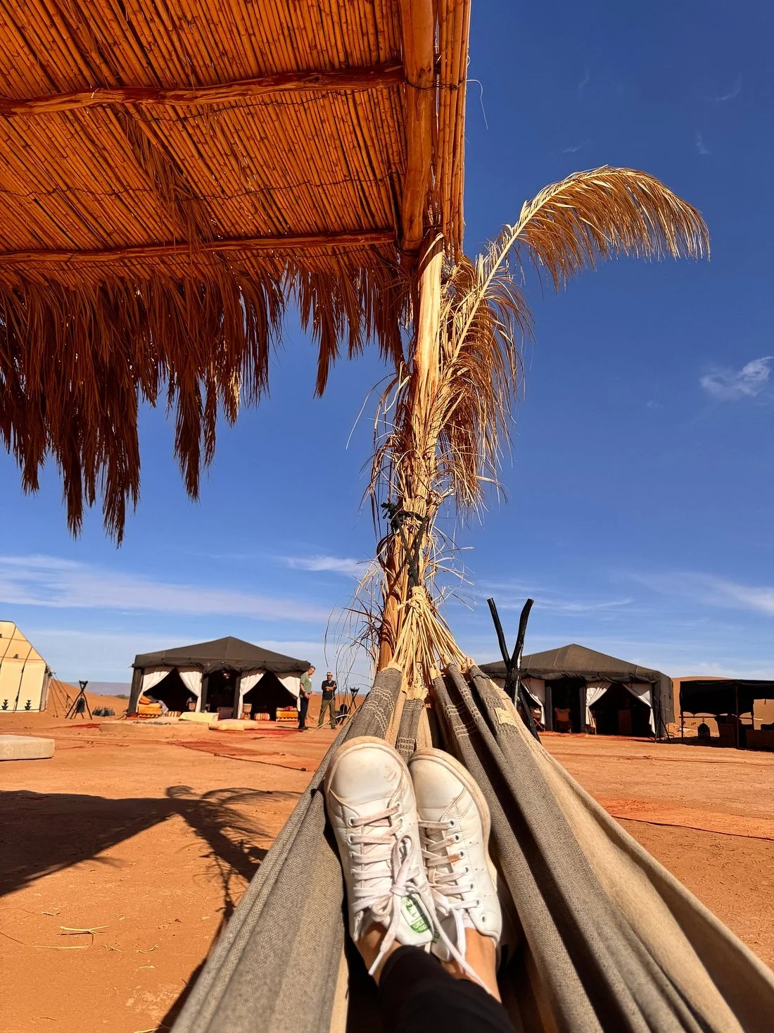 View from a hammock in a desert setting with a thatched roof overhead, several tents in the background, and a blue sky.