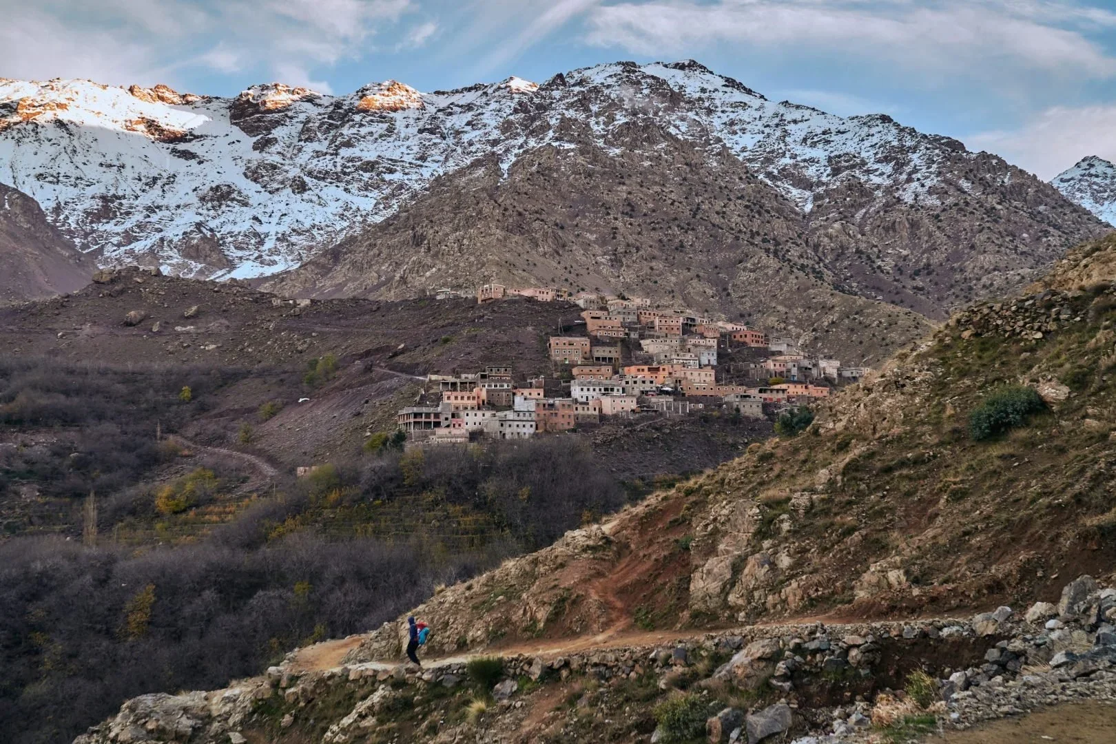 Mountain village with numerous buildings on a hillside, snow-capped peaks in the background, hikers on a trail in the foreground.
