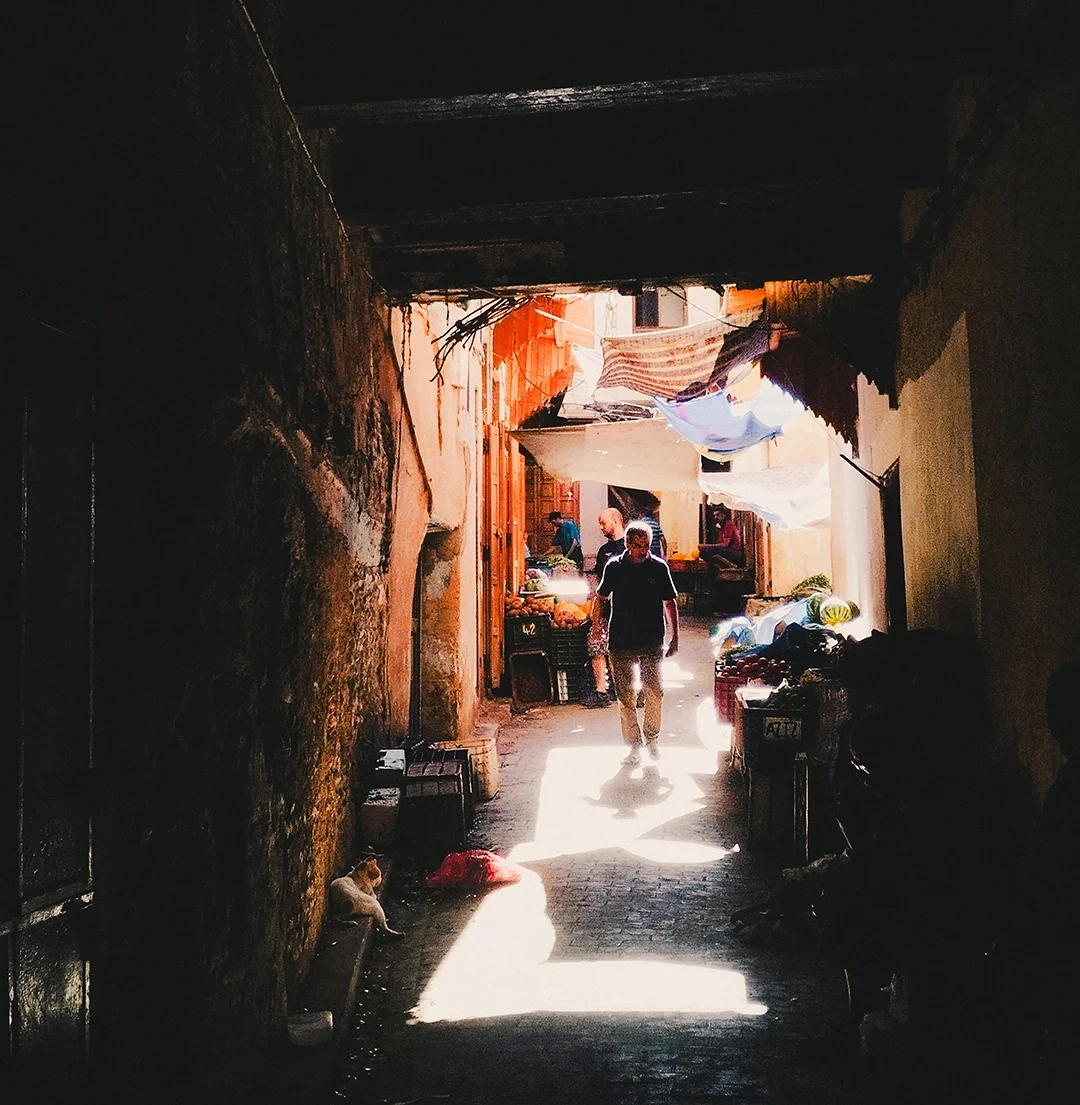 A narrow alleyway with a man walking toward the camera, illuminated by sunlight. The alley has hanging fabrics or coverings, with a small cat sitting on the ground to the left. In the background, there are market stalls and people.
