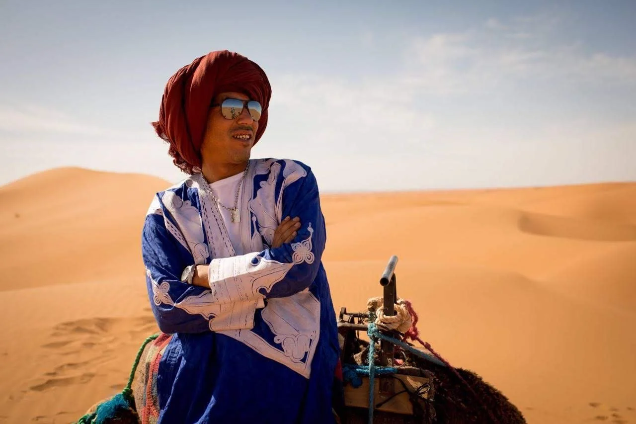 A man dressed in a blue and white flowing robe, wearing a red turban, sunglasses, and jewelry, stands in a desert landscape with sand dunes, crossing his arms and looking into the distance.