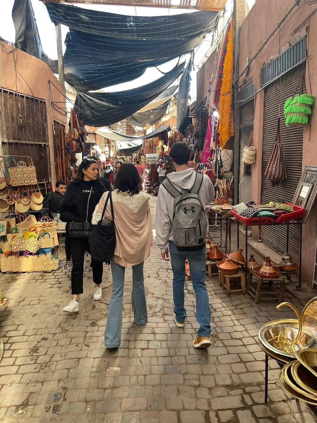 People shopping for handicrafts and accessories in an outdoor market with narrow cobblestone alleyways, covered by tarps.