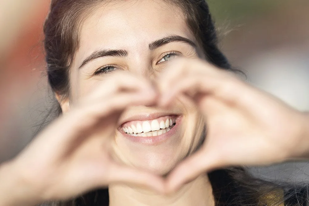 A smiling woman with light skin forming a heart shape with her hands.
