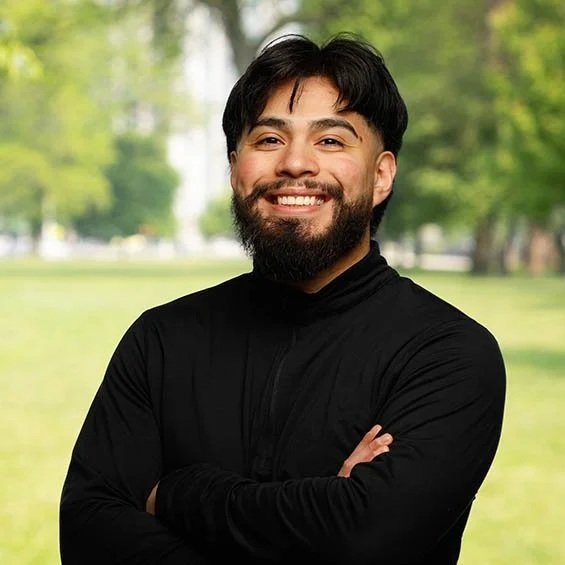 A smiling man with dark hair and a beard, wearing a black long-sleeve shirt, standing outdoors in a park.