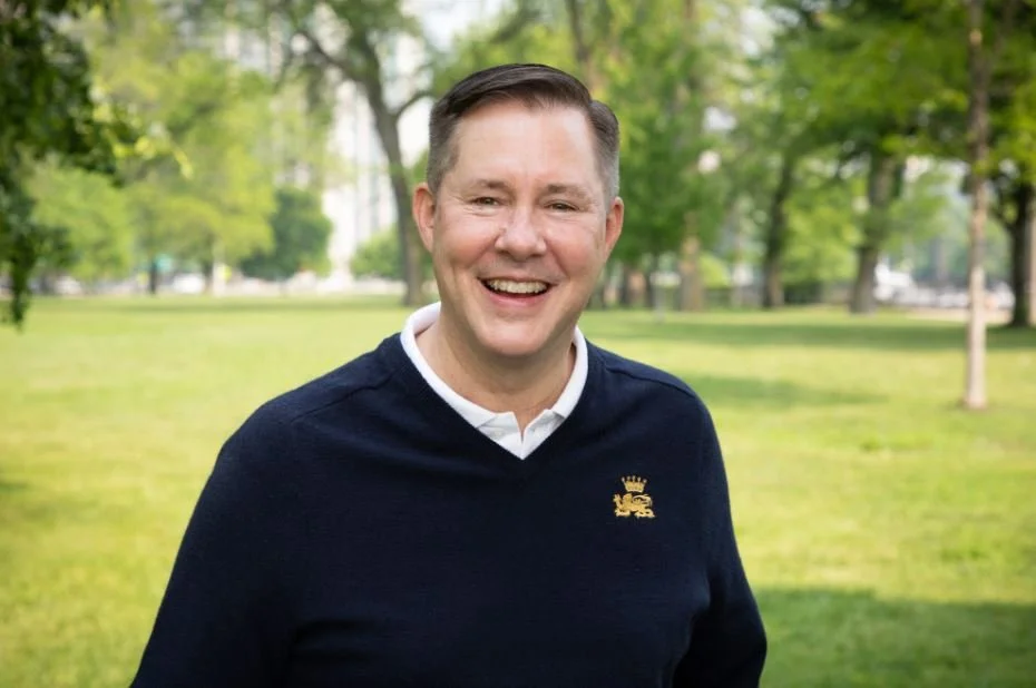 A smiling man in a navy blue sweater with a white collared shirt underneath, standing outdoors in a park with green grass and trees.