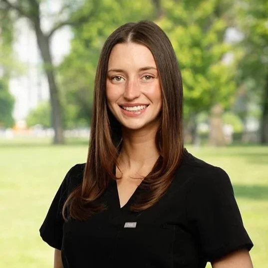 A young woman with long brown hair smiling outdoors in a park, wearing a black top.