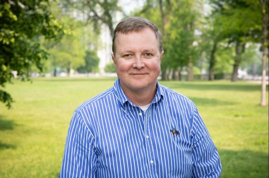 A man in a blue and white striped shirt standing in a park with green trees and grass in the background.