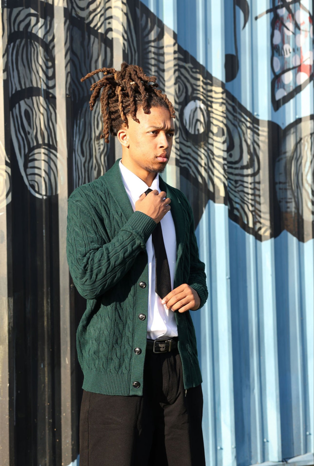 Young man with dreadlocks in a green cardigan, white shirt, and black tie standing in front of a wall featuring black, white, and blue graffiti art, looking serious and adjusting his tie.