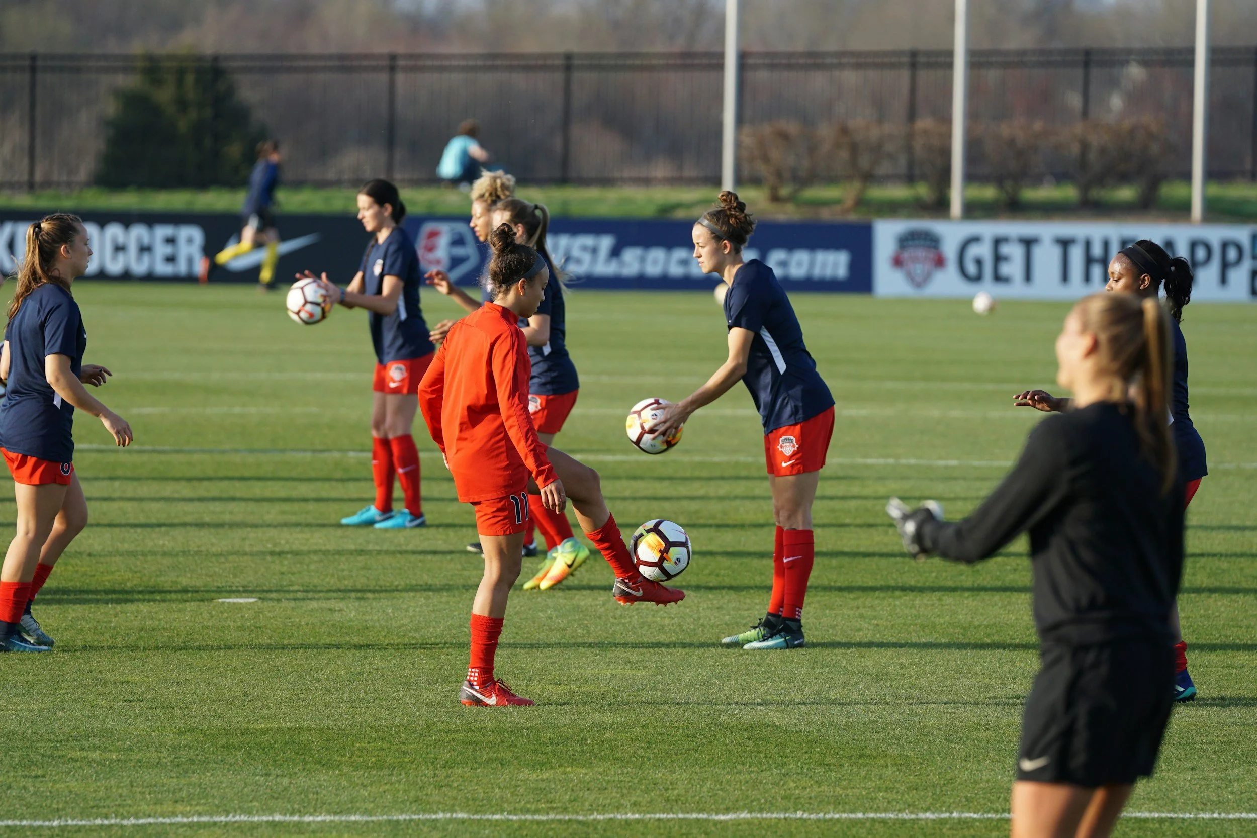 Women's soccer team practicing on the field with some players juggling soccer balls and others standing and waiting, wearing blue jerseys and red shorts, while a coach or player in black attire observes.